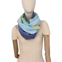 Mannequin wearing a hand-painted Otta pure linen scarf on a white background in 
blue, turquoise, green and purple tones. soft, fresh and lightweight, and elegant accessory measuring 70 x 190 cm. Unique artisan scarf with hand-sewn edges, perfect for women and men.
