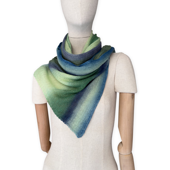 Mannequin wearing a colorful and lightweight linen scarf on a white background. Linen scarf is in shades of green and blue colors. It is hand-painted scarf in measures 35 x 202 cm by otta-italy scarves brand. Luxury Italian linen, soft and lightweight scarf for women and men.