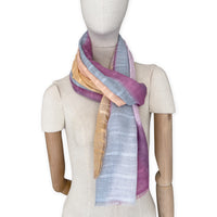 Mannequin wearing a hand-painted Otta pure linen scarf on a white background in 
pink, peach, fuchsia and gray tones. soft, fresh and lightweight, and elegant accessory measuring 70 x 190 cm. Unique artisan scarf with hand-sewn edges, perfect for women and men.
