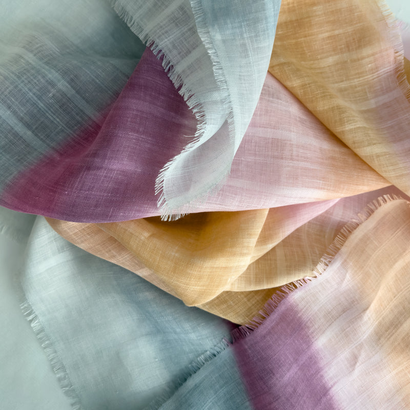 Close-up of soft fabric, a hand-painted Otta pure linen scarf draped softly, showcasing gentle shades of 
pink, peach, fuchsia and gray. Lightweight artisan accessory with delicate hand-sewn edges, measuring 70 x 190 cm.
