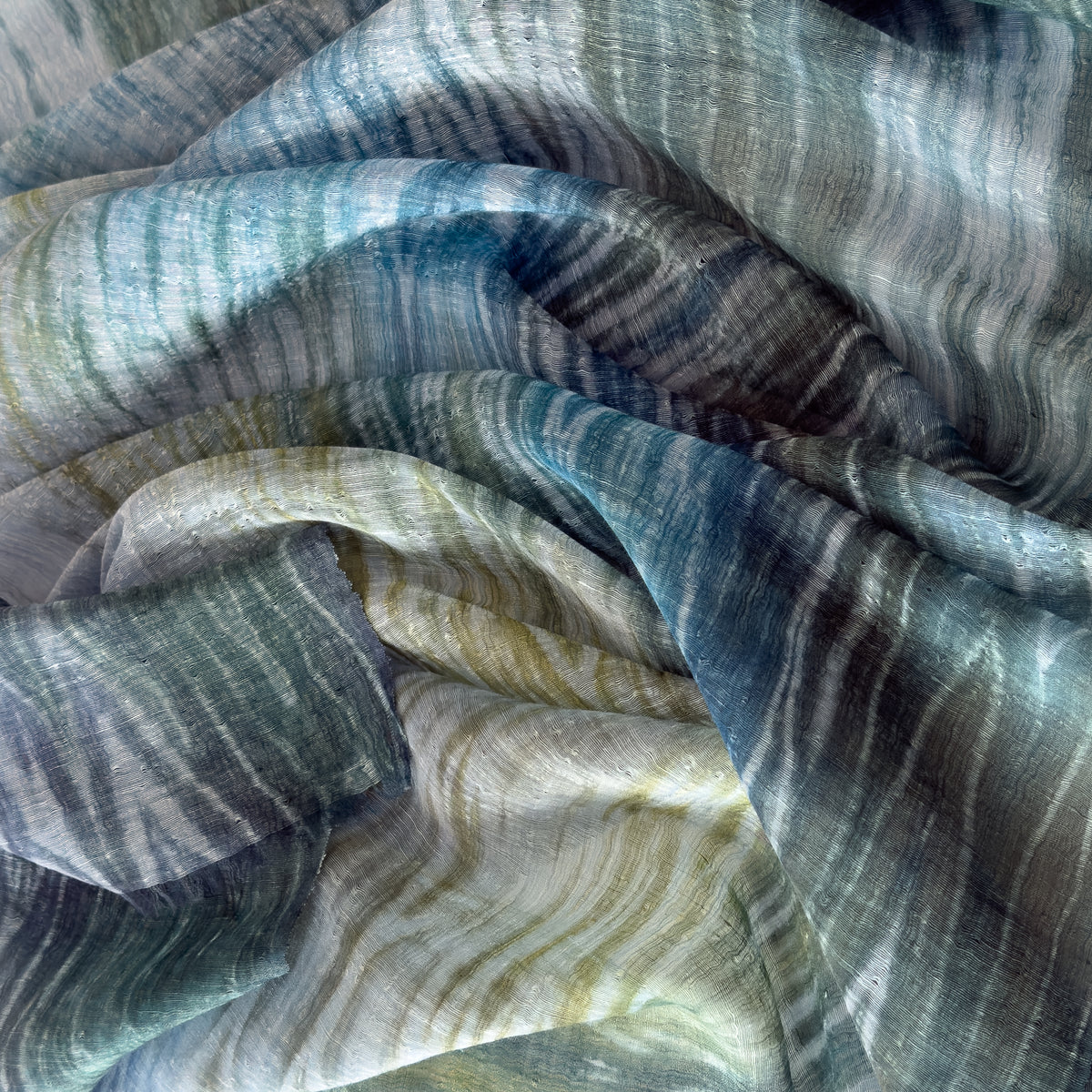 Close-up of soft fabric, a hand-painted Otta pure linen scarf draped softly, showcasing gentle shades of 
blue, light blue and green. Lightweight artisan accessory with delicate hand-sewn edges, measuring 52 x 205 cm.
