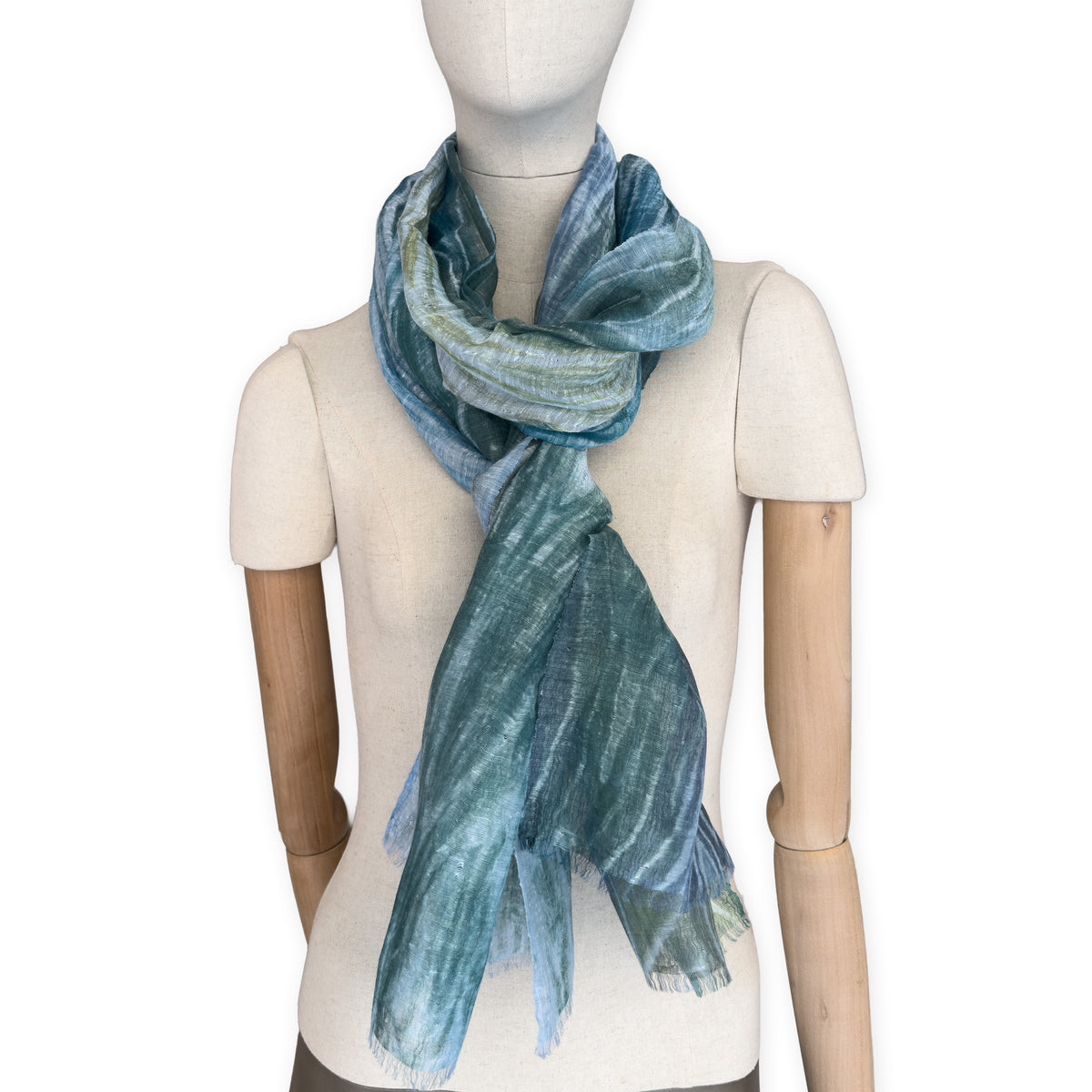 Mannequin wearing a hand-painted Otta pure linen scarf on a white background in 
blue, light blue and green tones. soft, fresh and lightweight, and elegant accessory measuring 52 x 205 cm. Unique artisan scarf with hand-sewn edges, perfect for women and men.
