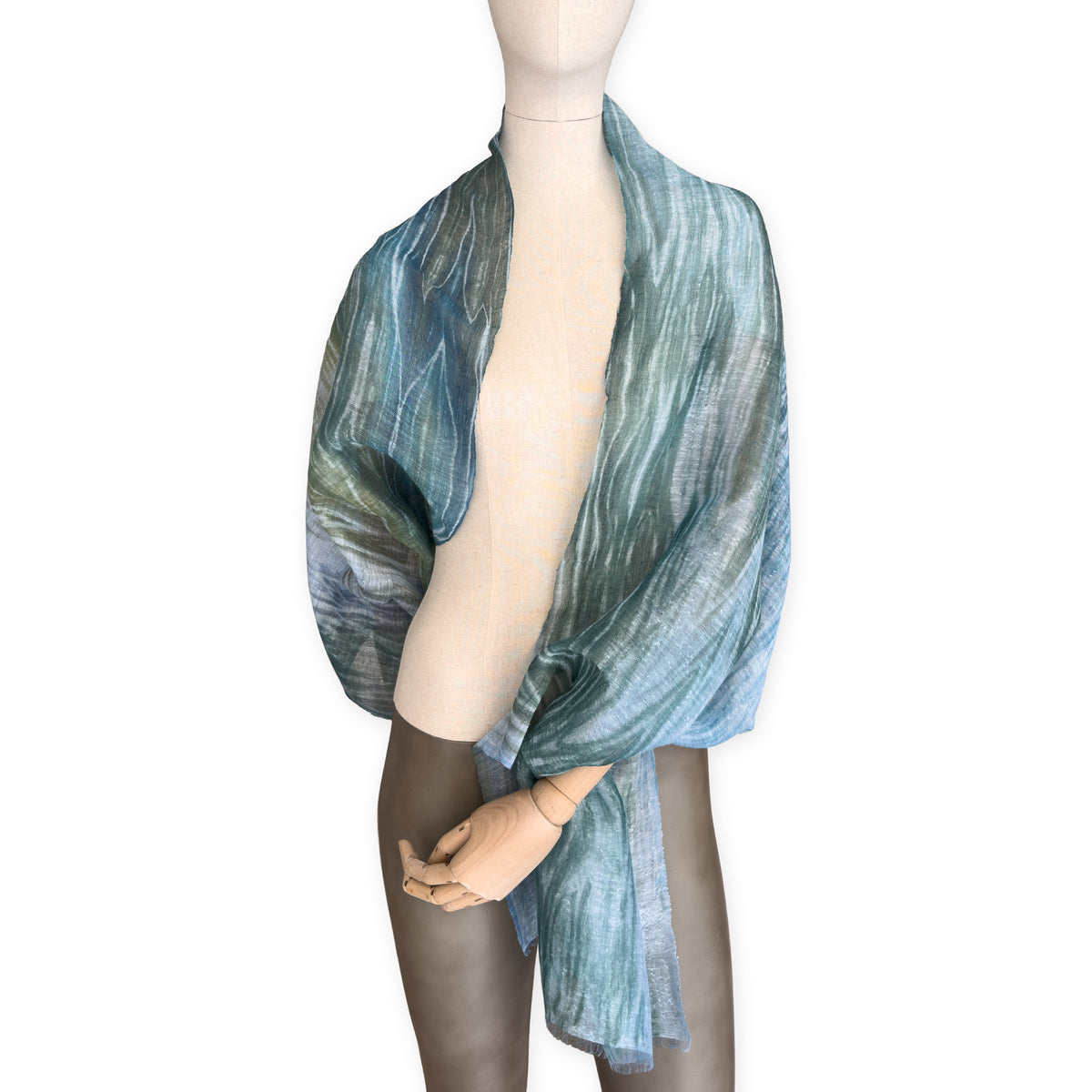 Mannequin wearing a hand-painted Otta pure linen scarf on a white background in 
blue, light blue and green tones. soft, fresh and lightweight, and elegant accessory measuring 52 x 205 cm. Unique artisan scarf with hand-sewn edges, perfect for women and men.
