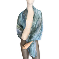 Mannequin wearing a hand-painted Otta pure linen scarf on a white background in 
blue, light blue and green tones. soft, fresh and lightweight, and elegant accessory measuring 52 x 205 cm. Unique artisan scarf with hand-sewn edges, perfect for women and men.
