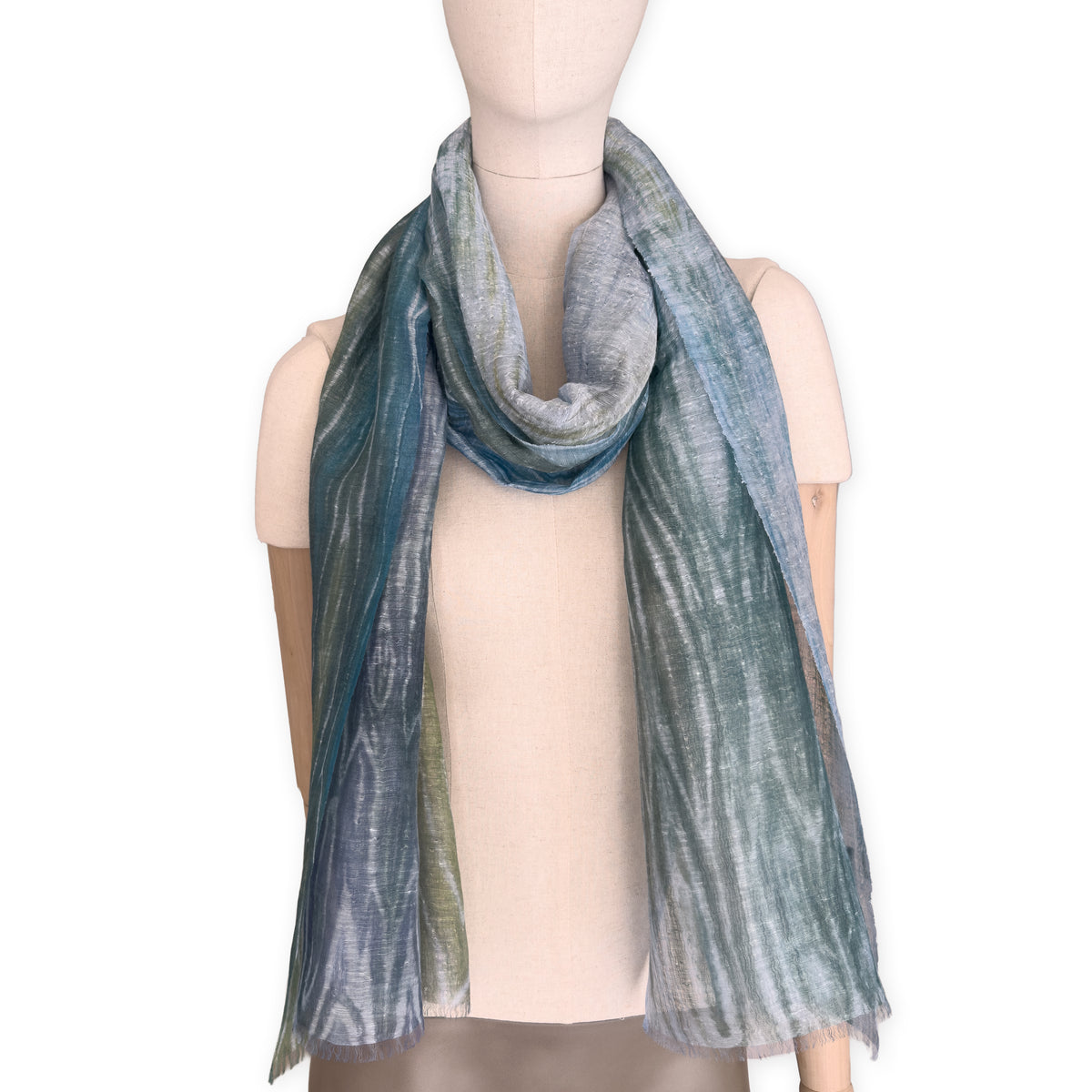 Mannequin wearing a hand-painted Otta pure linen scarf on a white background in 
blue, light blue and green tones. soft, fresh and lightweight, and elegant accessory measuring 52 x 205 cm. Unique artisan scarf with hand-sewn edges, perfect for women and men.

