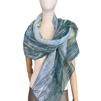 Mannequin wearing a hand-painted Otta pure linen scarf on a white background in 
blue, light blue and green tones. soft, fresh and lightweight, and elegant accessory measuring 52 x 205 cm. Unique artisan scarf with hand-sewn edges, perfect for women and men.
