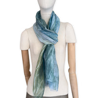 Mannequin wearing a hand-painted Otta pure linen scarf on a white background in 
blue, light blue and green tones. soft, fresh and lightweight, and elegant accessory measuring 52 x 205 cm. Unique artisan scarf with hand-sewn edges, perfect for women and men.
