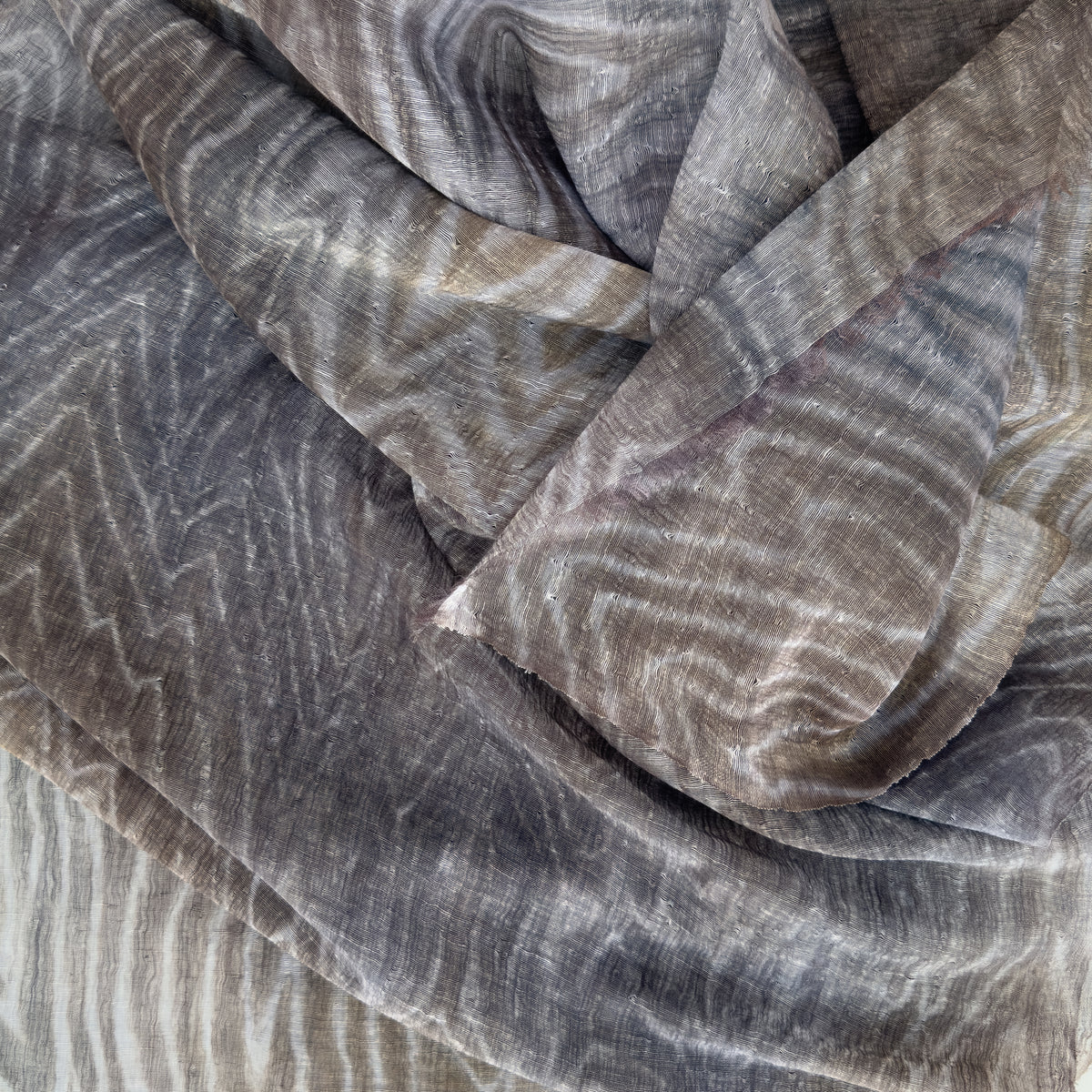 Close-up of soft fabric, a hand-painted Otta pure linen scarf draped softly, showcasing gentle shades of 
brown, gray and blue. Lightweight artisan accessory with delicate hand-sewn edges, measuring 51x 202 cm.
