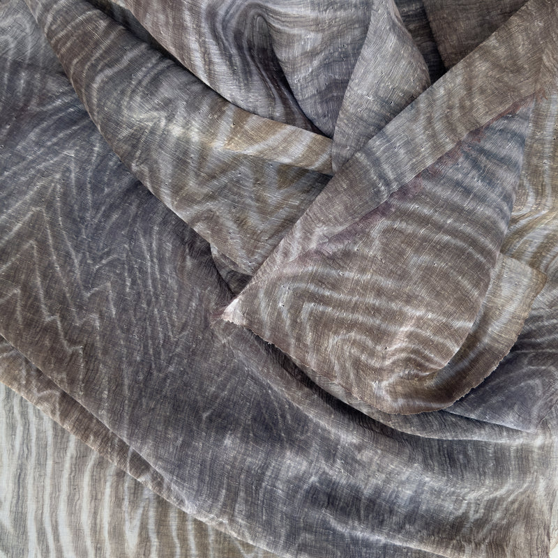 Close-up of soft fabric, a hand-painted Otta pure linen scarf draped softly, showcasing gentle shades of 
brown, gray and blue. Lightweight artisan accessory with delicate hand-sewn edges, measuring 51x 202 cm.
