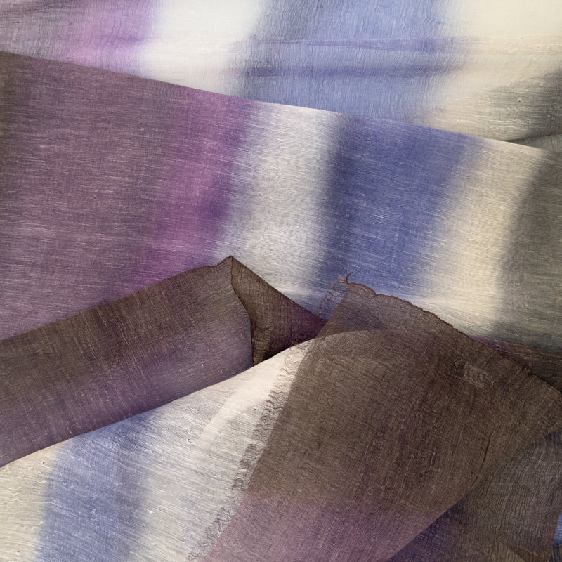 Close-up of a hand-painted Otta linen silk blend scarf showcasing soft shades of indigo, light blue, purple, brown and beige with delicate hand-fringed edges.
