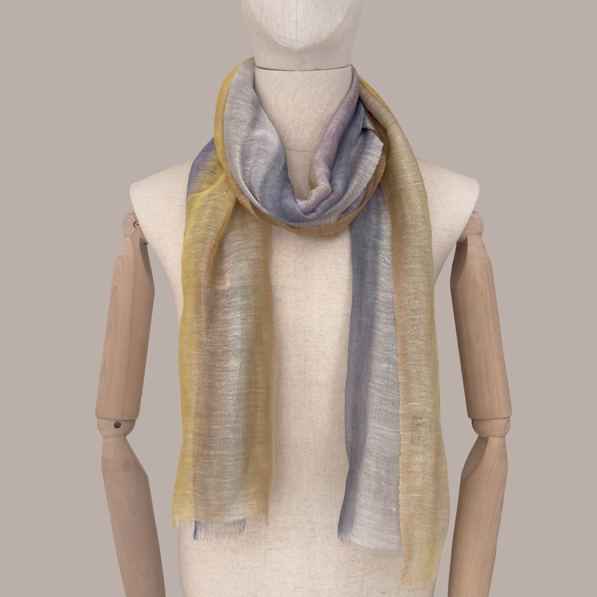 Mannequin wearing a hand-painted linen silk blend scarf by Otta-Italy, beige background. Yellow, ochre, sapphire, and gray wearable art. 52x200 cm.
