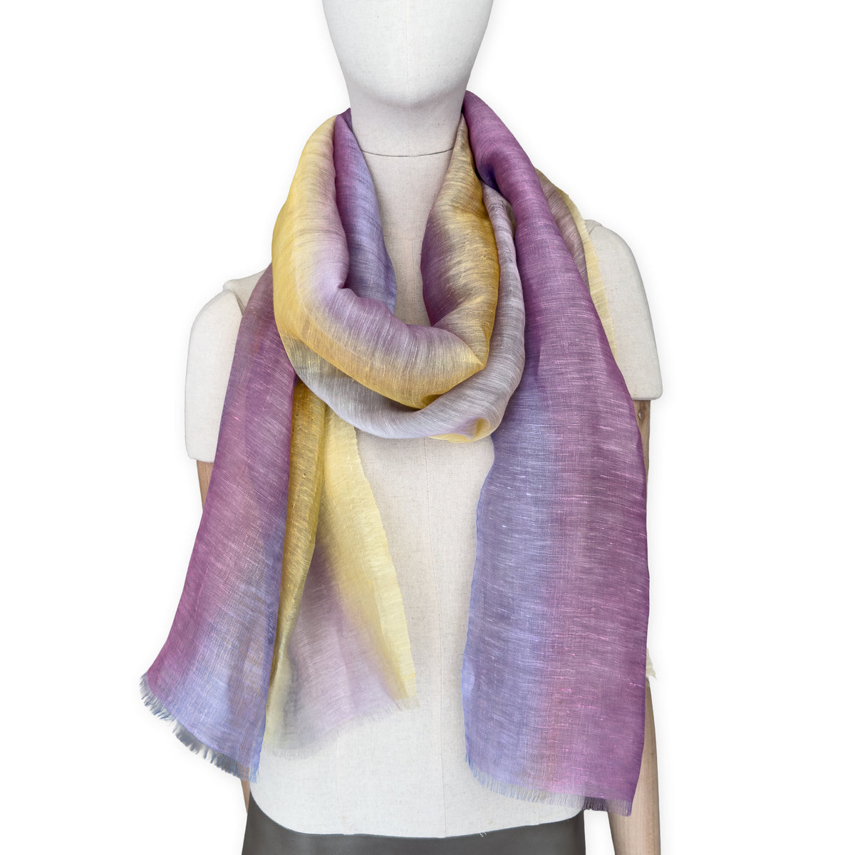 Mannequin wearing a hand-painted Otta pure linen scarf on a white background in 
yellow, purple and lilac tones. soft, fresh and lightweight, and elegant accessory measuring 52 x 200 cm. Unique artisan scarf with hand-sewn edges, perfect for women and men.
