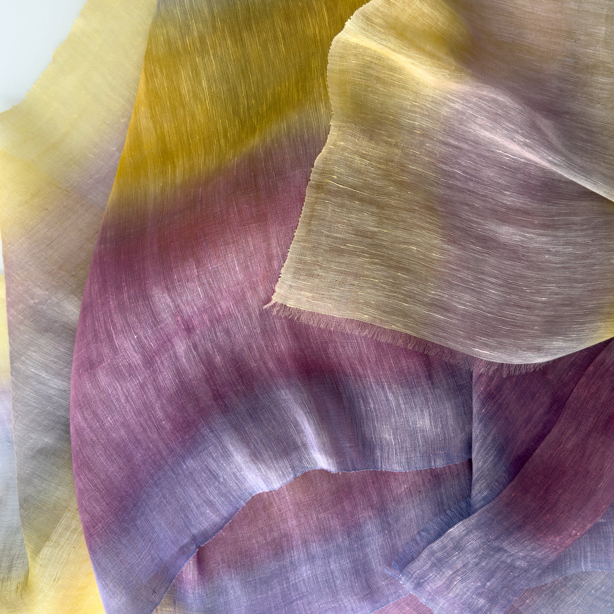 Close-up of soft fabric, a hand-painted Otta pure linen scarf draped softly, showcasing gentle shades of 
yellow, purple and lilac. Lightweight artisan accessory with delicate hand-sewn edges, measuring 52 x 200 cm.

