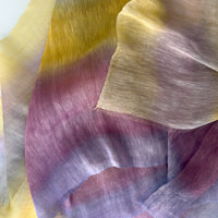 Close-up of soft fabric, a hand-painted Otta pure linen scarf draped softly, showcasing gentle shades of 
yellow, purple and lilac. Lightweight artisan accessory with delicate hand-sewn edges, measuring 52 x 200 cm.
