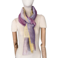 Mannequin wearing a hand-painted Otta pure linen scarf on a white background in 
yellow, purple and lilac tones. soft, fresh and lightweight, and elegant accessory measuring 52 x 200 cm. Unique artisan scarf with hand-sewn edges, perfect for women and men.
