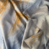 Close-up of a hand-painted Otta silk cotton blend scarf draped softly, showcasing gentle shades of 
blue, gray and orange. Lightweight artisan accessory with delicate hand-fringed edges, measuring 67 x 193 cm.
