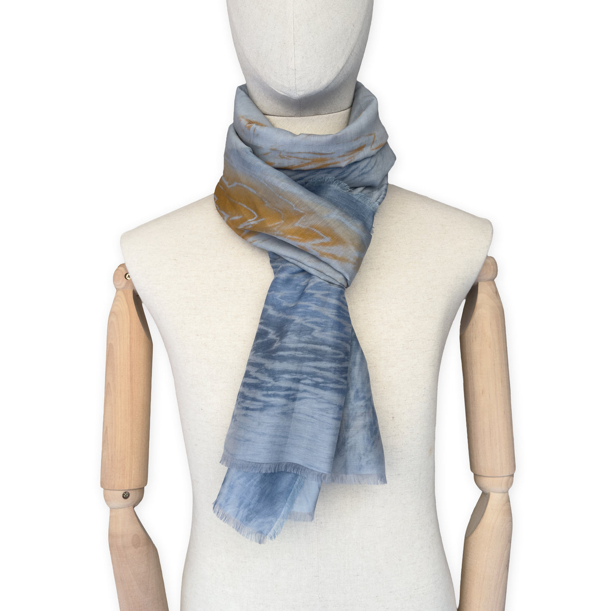 Mannequin wearing a hand-painted Otta silk cotton blend scarf on a white background in 
blue, gray and orange e tones. soft, fresh and lightweight, and elegant accessory measuring 67 x 193 cm. Unique artisan scarf with hand-fringed edges, perfect for women and men.
