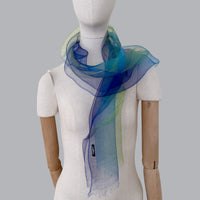 Mannequin wearing a hand-painted pure silk organza scarf by Otta-Italy, gray background. Blue, green and violet. 45x180 cm.
