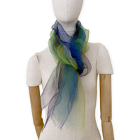 Mannequin wearing an elegant hand-painted Otta pure silk organza scarf on a gray background. Blue, green and violet tones.
