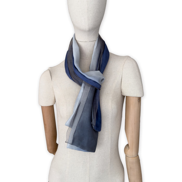 Mannequin wearing a colorful and lightweight silk scarf on a white background. Silk scarf is in shades of gray and midnight blue colors. It is hand-painted scarf in measures 24 x 168 cm by otta-italy scarves brand.
