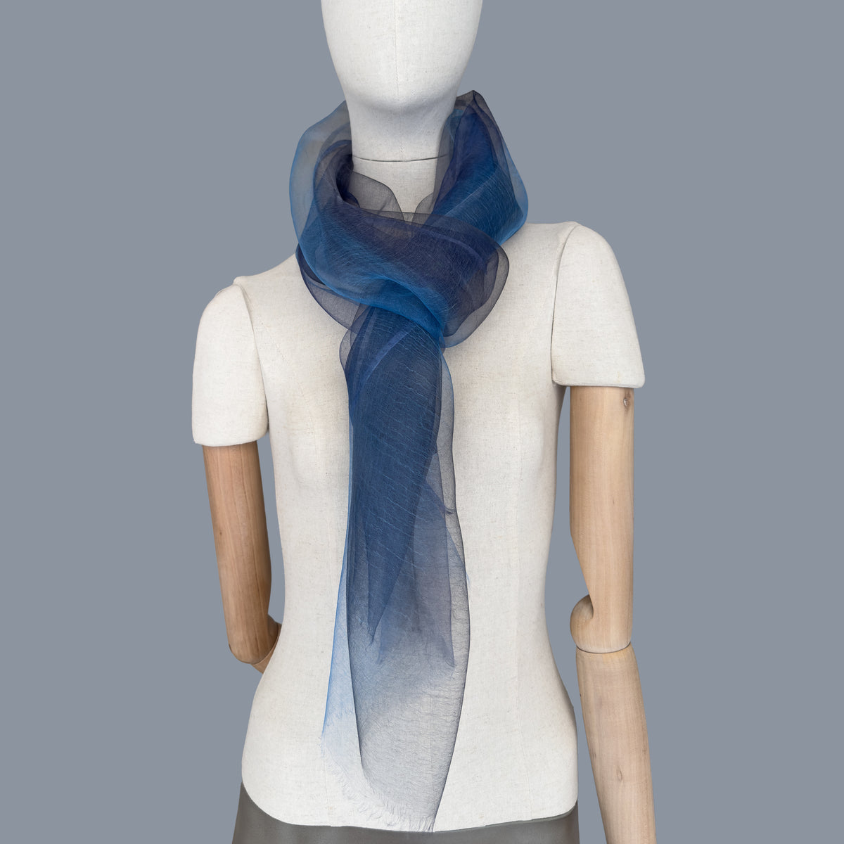 Mannequin wearing a colorful and lightweight silk scarf on a gray background. Silk scarf is in shades of gray, midnight blue and blue colors. It is hand-painted scarf in measures 50 x 200 cm by otta-italy scarves brand.
