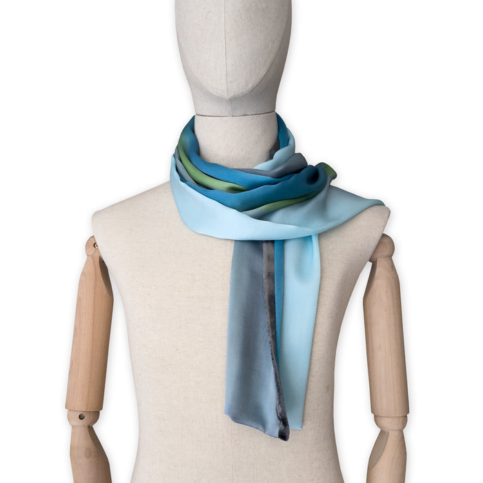 Mannequin wearing a colorful and lightweight silk scarf on a white background. Silk scarf is in shades of gray, light blue, turquoise and green colors. It is hand-painted scarf in measures 172 x 22 cm by otta-italy scarves brand.

