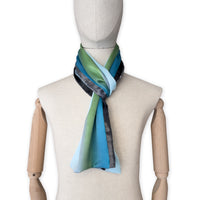 Mannequin wearing a colorful and lightweight silk scarf on a white background. Silk scarf is in shades of gray, light blue, turquoise and green colors. It is hand-painted scarf in measures 172 x 22 cm by otta-italy scarves brand.
