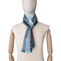 Mannequin wearing a colorful and lightweight silk scarf on a white background. Silk scarf is in shades of gray, light blue, turquoise and green colors. It is hand-painted scarf in measures 172 x 22 cm by otta-italy scarves brand.
