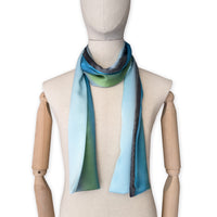 Mannequin wearing a colorful and lightweight silk scarf on a white background. Silk scarf is in shades of gray, light blue, turquoise and green colors. It is hand-painted scarf in measures 172 x 22 cm by otta-italy scarves brand.
