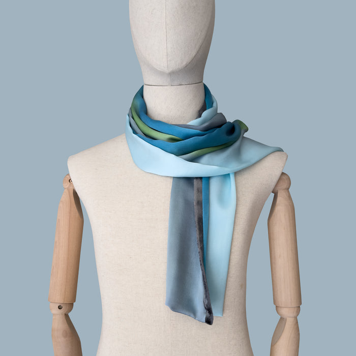 Mannequin wearing a colorful and lightweight silk scarf on a blue background. Silk scarf is in shades of gray, light blue, turquoise and green colors. It is hand-painted scarf in measures 172 x 22 cm by otta-italy scarves brand.
