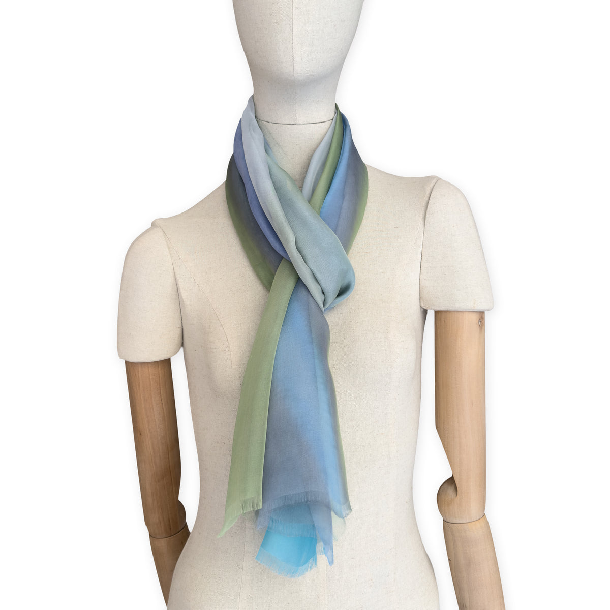 Mannequin wearing a hand-painted Otta pure silk scarf on a white background in 
blue, green and gray tones. soft, fresh and lightweight, and elegant accessory measuring 65 x 175 cm. Unique artisan scarf with hand-fringed edges, perfect for women and men.
