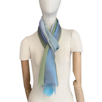 Mannequin wearing a hand-painted Otta pure silk scarf on a white background in 
blue, green and gray tones. soft, fresh and lightweight, and elegant accessory measuring 65 x 175 cm. Unique artisan scarf with hand-fringed edges, perfect for women and men.
