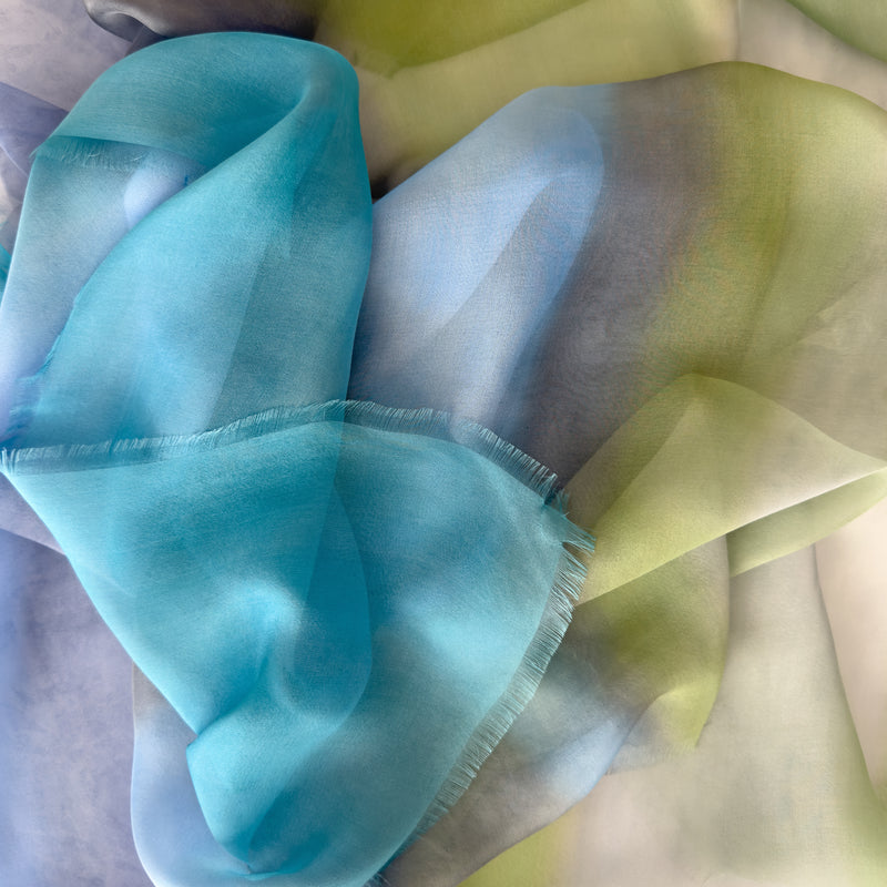 Close-up of a hand-painted Otta pure silk scarf draped softly, showcasing gentle shades of 
blue, green and gray. Lightweight artisan accessory with delicate hand-fringed edges, measuring 65 x 175 cm.
