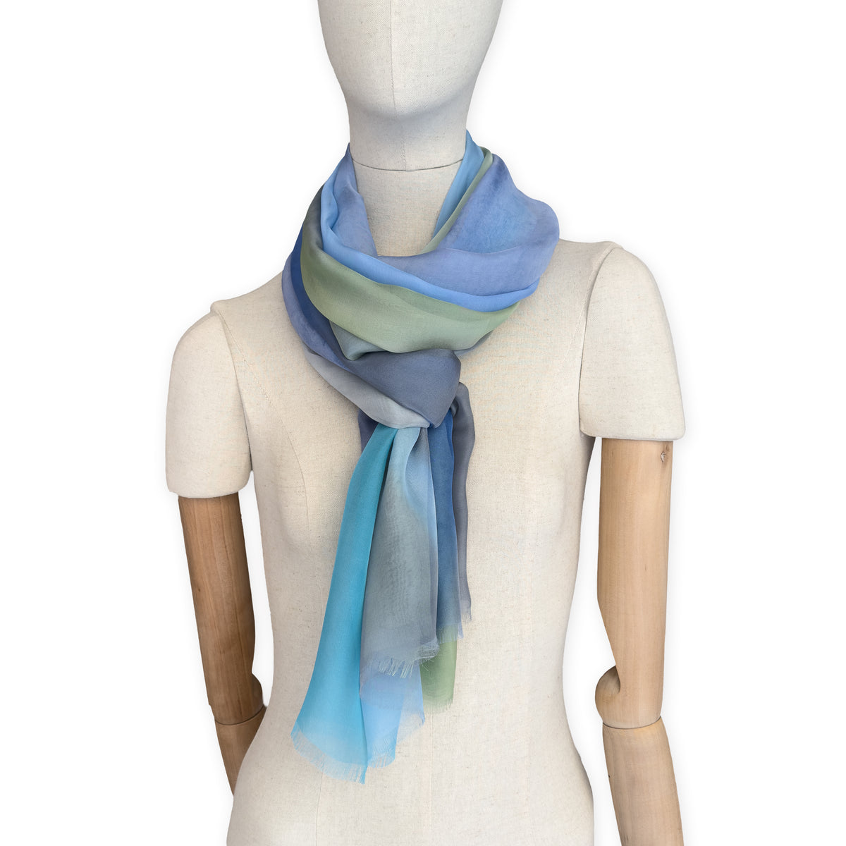 Mannequin wearing a hand-painted Otta pure silk scarf on a white background in 
blue, green and gray tones. soft, fresh and lightweight, and elegant accessory measuring 65 x 175 cm. Unique artisan scarf with hand-fringed edges, perfect for women and men.
