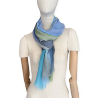 Mannequin wearing a hand-painted Otta pure silk scarf on a white background in 
blue, green and gray tones. soft, fresh and lightweight, and elegant accessory measuring 65 x 175 cm. Unique artisan scarf with hand-fringed edges, perfect for women and men.
