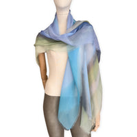 Mannequin wearing a hand-painted Otta pure silk scarf on a white background in 
blue, green and gray tones. soft, fresh and lightweight, and elegant accessory measuring 65 x 175 cm. Unique artisan scarf with hand-fringed edges, perfect for women and men.
