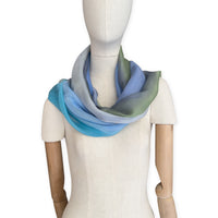 Mannequin wearing a hand-painted Otta pure silk scarf on a white background in 
blue, green and gray tones. soft, fresh and lightweight, and elegant accessory measuring 65 x 175 cm. Unique artisan scarf with hand-fringed edges, perfect for women and men.
