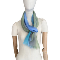 Mannequin wearing a hand-painted Otta pure silk scarf on a white background in 
blue, green and gray tones. soft, fresh and lightweight, and elegant accessory measuring 65 x 175 cm. Unique artisan scarf with hand-fringed edges, perfect for women and men.
