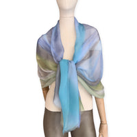 Mannequin wearing a hand-painted Otta pure silk scarf on a white background in 
blue, green and gray tones. soft, fresh and lightweight, and elegant accessory measuring 65 x 175 cm. Unique artisan scarf with hand-fringed edges, perfect for women and men.
