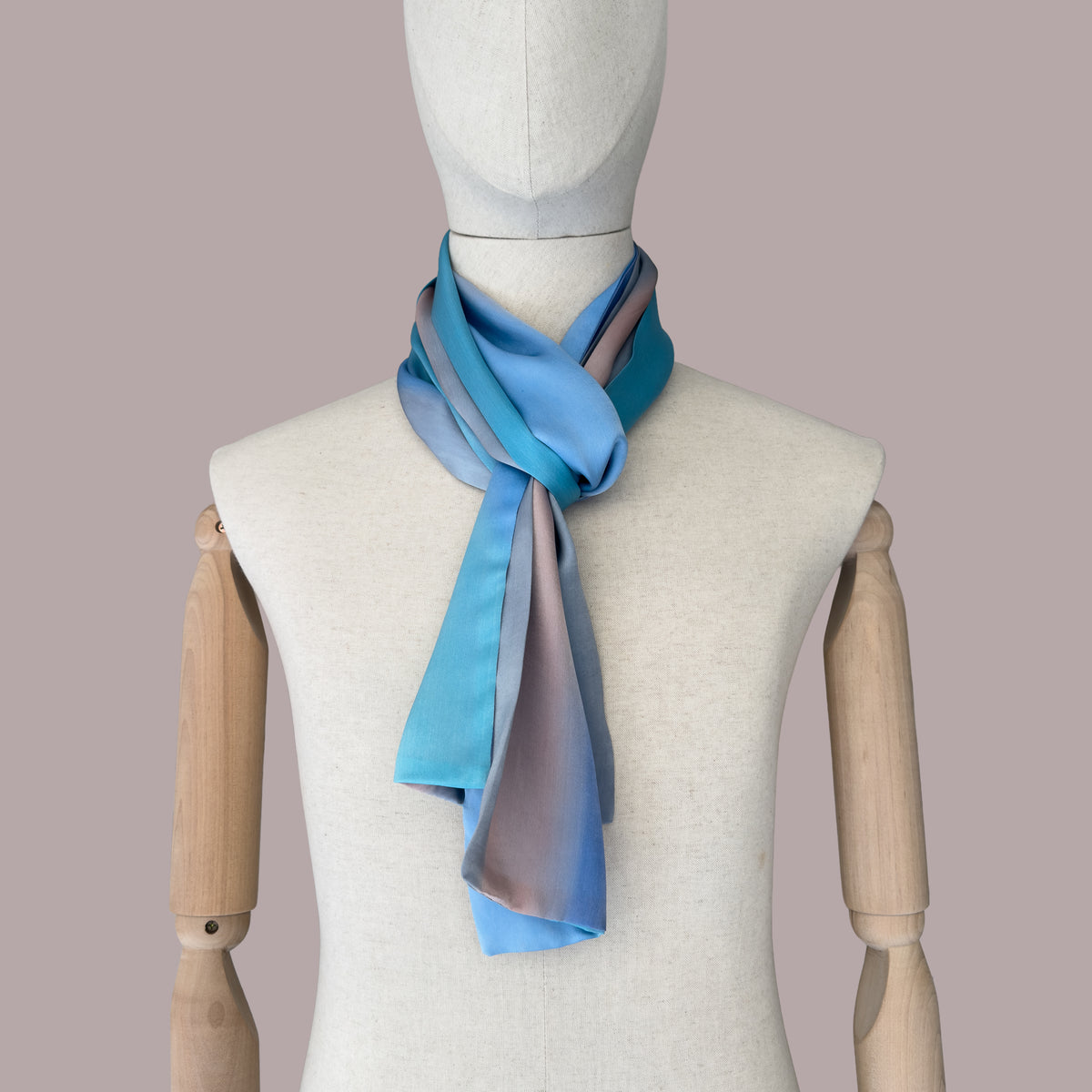 Mannequin wearing scarf on a beige background. Hand-painted silk scarf by Otta-Italy, 45 x 180 cm, featuring 
indigo, turquoise, sky blue, gray and peach. Luxury Italian pure silk accessory, soft and lightweight scarf for women and men.
