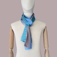 Mannequin wearing scarf on a beige background. Hand-painted silk scarf by Otta-Italy, 45 x 180 cm, featuring 
indigo, turquoise, sky blue, gray and peach. Luxury Italian pure silk accessory, soft and lightweight scarf for women and men.
