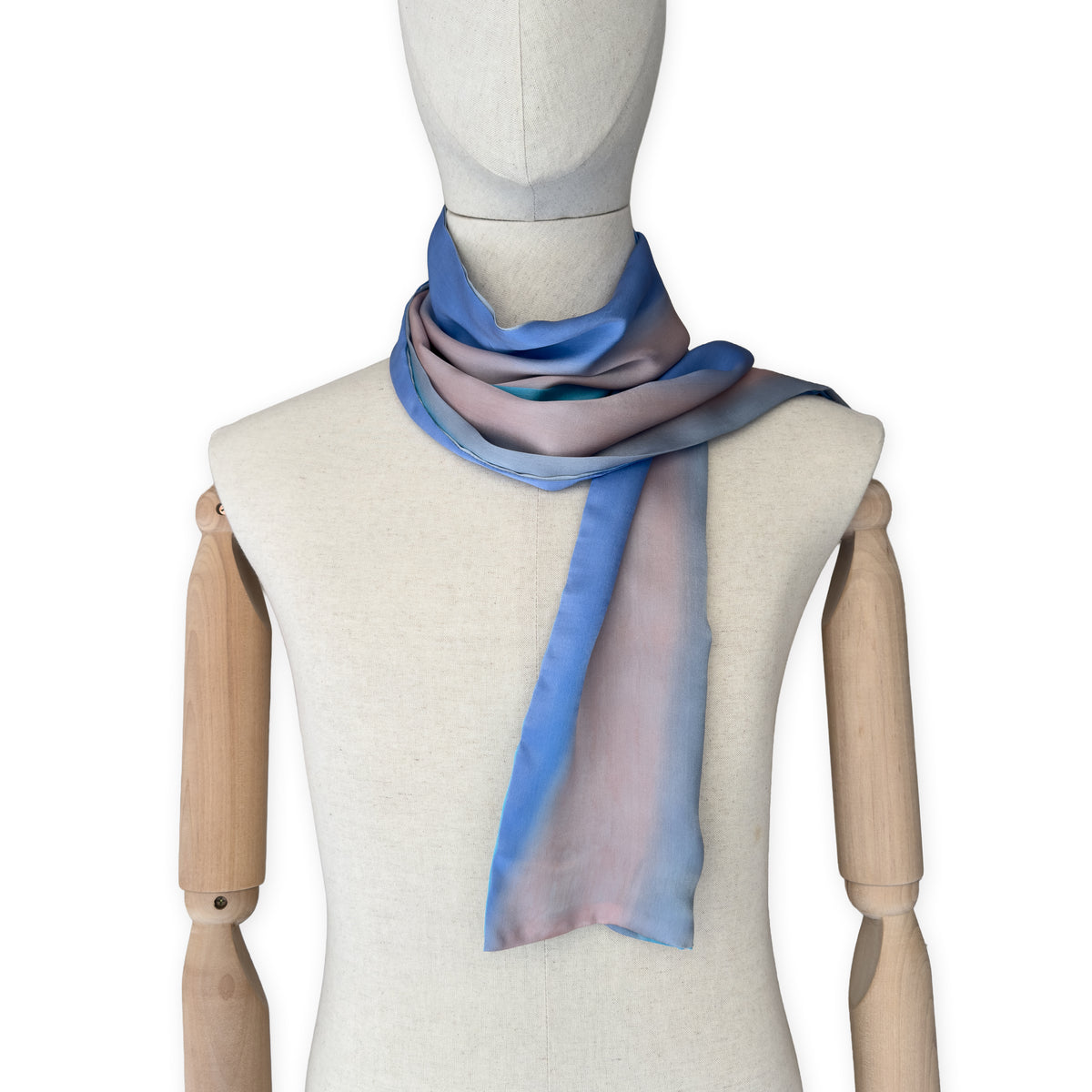 Mannequin wearing a hand-painted Otta silk scarf on a white background in 
indigo, turquoise, sky blue, gray and peach tones. Lightweight, soft, and elegant accessory measuring 45 x 180 cm. Unique artisan scarf with hand-hem edges, perfect for women and men.
