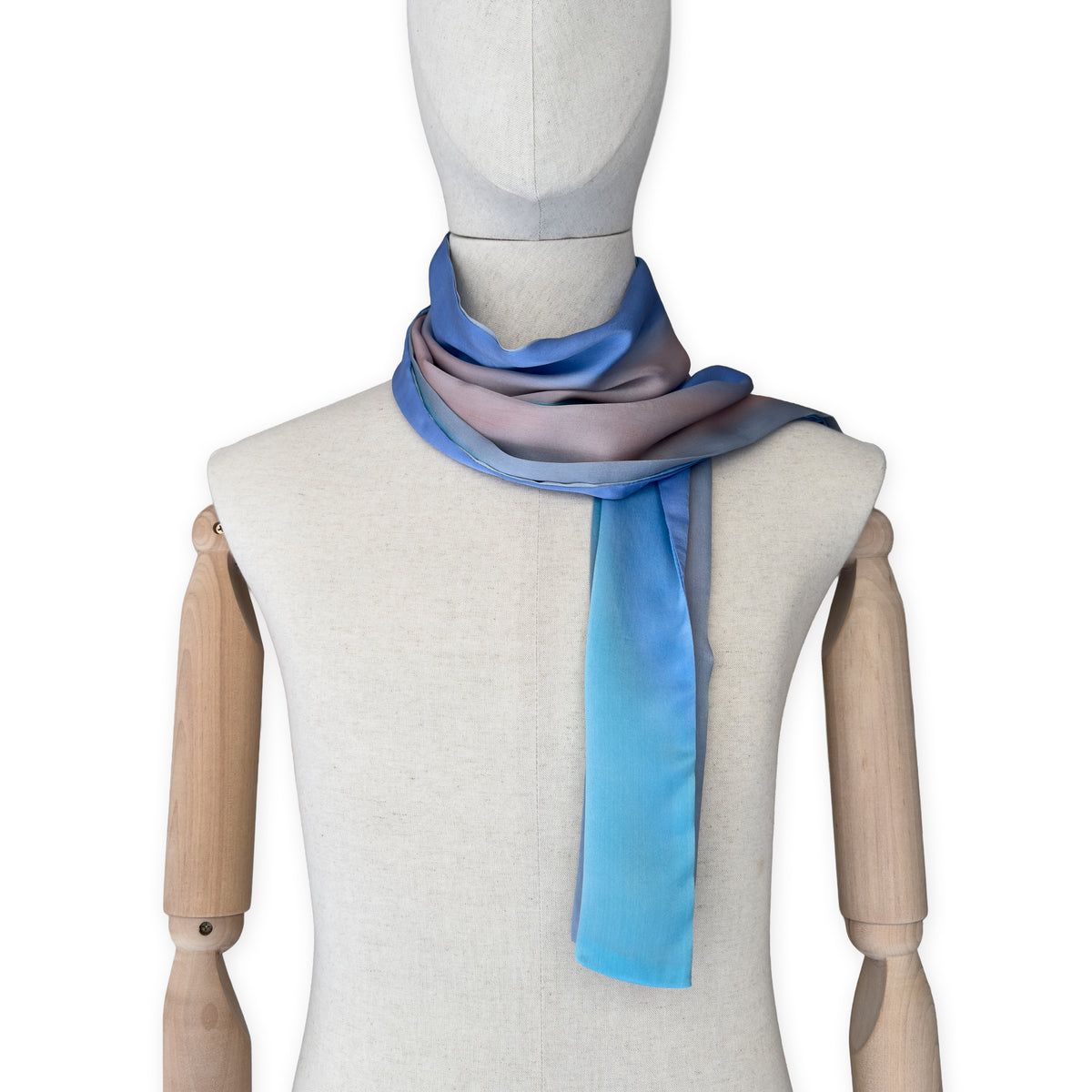 Mannequin wearing a hand-painted Otta silk scarf on a white background in 
indigo, turquoise, sky blue, gray and peach tones. Lightweight, soft, and elegant accessory measuring 45 x 180 cm. Unique artisan scarf with hand-hem edges, perfect for women and men.
