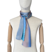 Mannequin wearing a hand-painted Otta silk scarf on a white background in 
indigo, turquoise, sky blue, gray and peach tones. Lightweight, soft, and elegant accessory measuring 45 x 180 cm. Unique artisan scarf with hand-hem edges, perfect for women and men.
