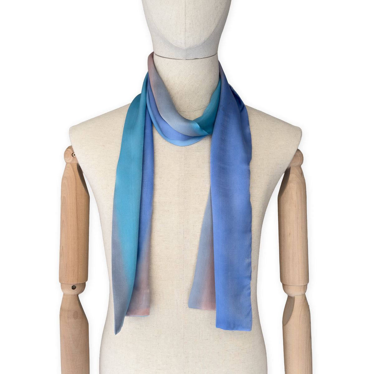 Mannequin wearing a hand-painted Otta silk scarf on a white background in 
indigo, turquoise, sky blue, gray and peach tones. Lightweight, soft, and elegant accessory measuring 45 x 180 cm. Unique artisan scarf with hand-hem edges, perfect for women and men.
