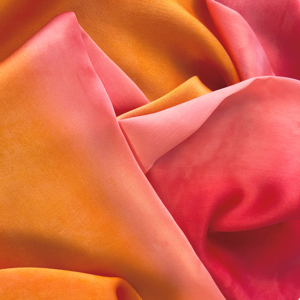 Close-up of a hand-painted Otta silk scarf draped softly, showcasing gentle shades of 
pink, fuchsia and orange. Lightweight artisan accessory with delicate hand-hem, measuring 22 x 170 cm.
