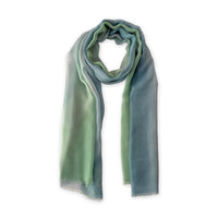 A hand-painted Otta pure silk scarf in 
green and blue, loosely looped to showcase its full length and delicate texture in white background. Lightweight artisan accessory with hand-fringed edges, measuring 65 x 175 cm.
