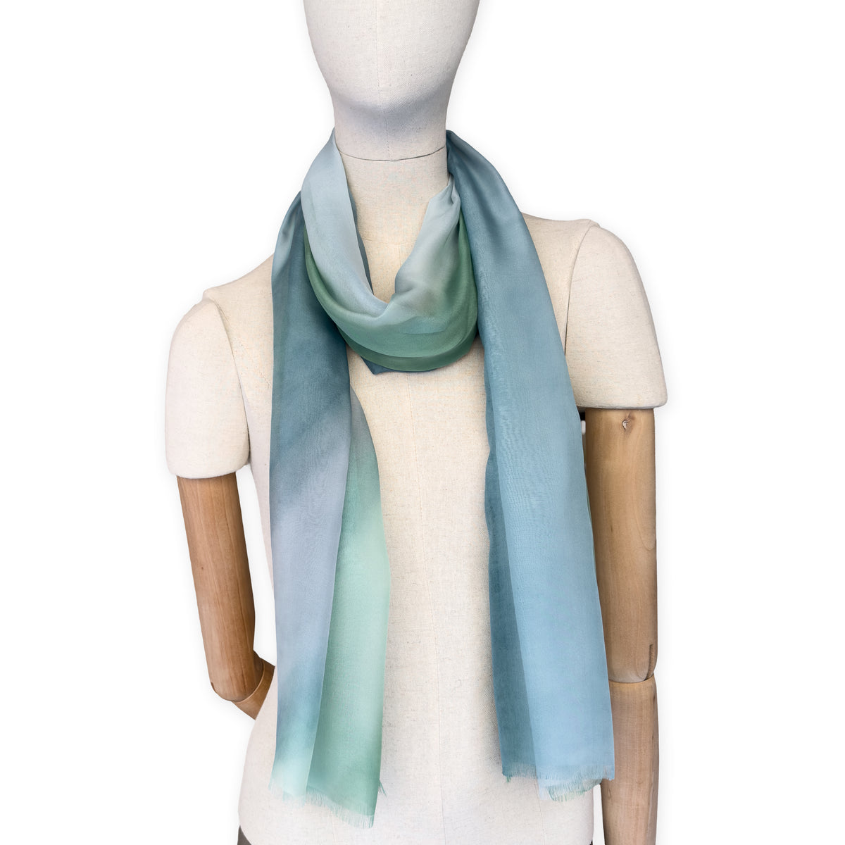 Mannequin wearing a hand-painted Otta pure silk scarf on a white background in 
green and blue tones. soft, fresh and lightweight, and elegant accessory measuring 65 x 175 cm. Unique artisan scarf with hand-fringed edges, perfect for women and men.
