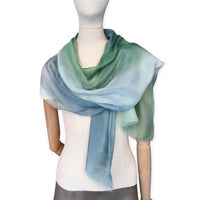 Mannequin wearing a hand-painted Otta pure silk scarf on a white background in 
green and blue tones. soft, fresh and lightweight, and elegant accessory measuring 65 x 175 cm. Unique artisan scarf with hand-fringed edges, perfect for women and men.
