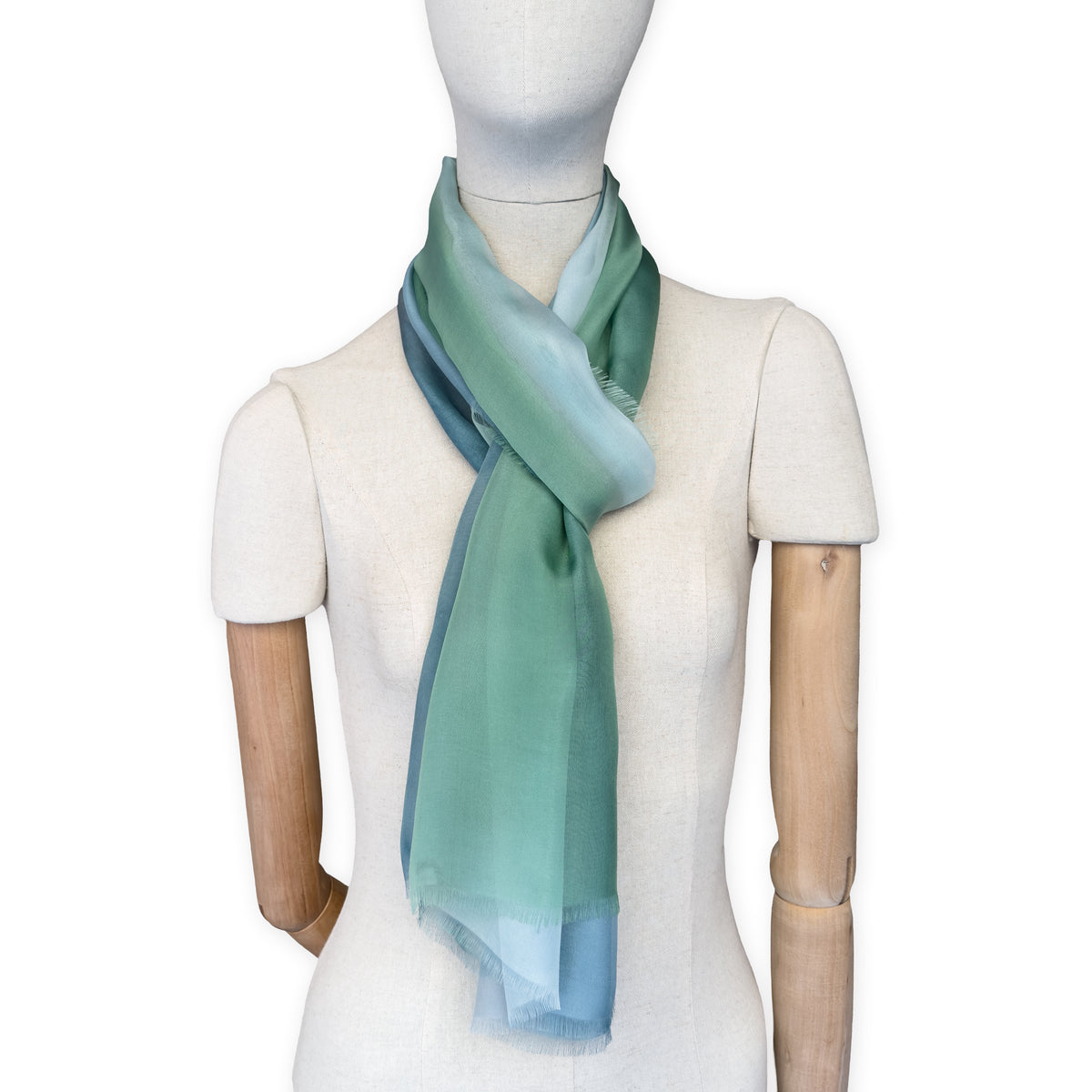Mannequin wearing a hand-painted Otta pure silk scarf on a white background in 
green and blue tones. soft, fresh and lightweight, and elegant accessory measuring 65 x 175 cm. Unique artisan scarf with hand-fringed edges, perfect for women and men.
